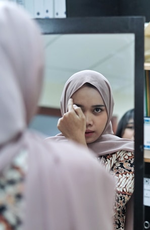 A woman wearing a hijab is looking at herself in a mirror, adjusting her hijab with one hand. The reflection captures her focused expression. She is in an indoor setting, possibly an office or a home, as indicated by the shelves and files in the background.