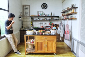 A cozy kitchen with a wooden island in the center topped with fruit baskets and various kitchen items. Shelves filled with jars of spices line the walls, and aprons hang neatly on hooks. A man stands by the window, sipping from a cup, creating a relaxed atmosphere. A variety of kitchen utensils and supplies are organized neatly, with a clock positioned above a tiled backsplash.