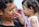 A young child in a pink outfit is gently reaching towards the face of a smiling adult. The adult holds the child close, and there is a sense of affection and bonding between them. The child has curly hair and is wearing an earring, while the adult has short, curly hair.