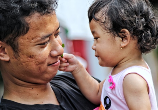 A young child in a pink outfit is gently reaching towards the face of a smiling adult. The adult holds the child close, and there is a sense of affection and bonding between them. The child has curly hair and is wearing an earring, while the adult has short, curly hair.