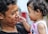 A young child in a pink outfit is gently reaching towards the face of a smiling adult. The adult holds the child close, and there is a sense of affection and bonding between them. The child has curly hair and is wearing an earring, while the adult has short, curly hair.