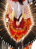 Elaborate feathered headdress with a symmetrical design featuring dark brown feathers edged with white spots. Bright red, yellow, and orange patterns form a circular design at the center, with additional fringe details in the background.