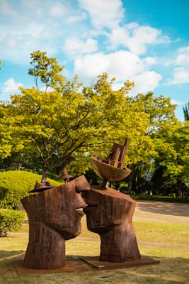 Two large sculptures depicting human faces in profile are positioned close to each other with additional abstract elements perched on top. The sculptures are made of a weathered, rusted metal, set against a background of vibrant green trees under a bright blue sky with fluffy white clouds.