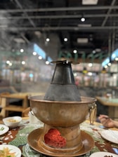 A traditional hot pot set on a dining table, with steam rising from the pot, indicating warmth and cooking activity. The table is adorned with various plates and bowls containing food. The background displays a blurred view of a restaurant setting with dim lighting and hanging decorations.