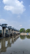 A series of concrete structures with metal railings extends across a body of water, likely a dam or floodgate system. The sky is partly cloudy, and trees are visible in the background. The water reflects both the structures and the surrounding environment.