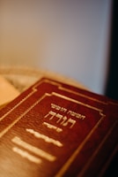 A close-up view of a book with a red cover featuring gold embossed Hebrew text. The book is positioned at an angle, with soft lighting creating a warm ambiance.