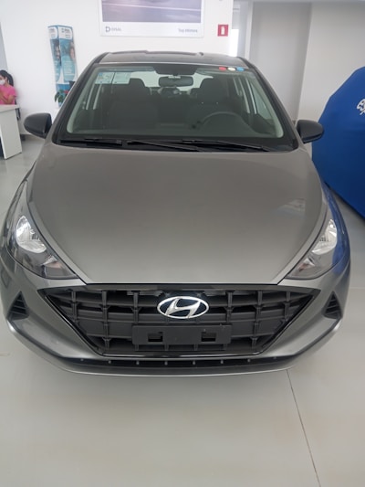 A grey Hyundai vehicle is displayed indoors on a smooth showroom floor. The front grille features a prominent Hyundai logo, and the vehicle's headlights have a sleek design. In the background, there is a partial view of a person seated and some promotional materials.