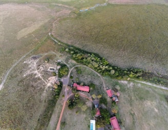 The aerial view depicts rural areas with sparse vegetation and scattered buildings with red roofs. Visible are a few dirt paths and roads, a swimming pool, and some parked vehicles. The landscape is a mix of grassy fields, patches of trees, and scrubland. The scene appears calm and untouched by urban development.