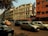 A busy street scene featuring several cars on a road with a distinctive yellow taxi. The backdrop consists of a blend of colonial and modern architecture, with a tall, ornate beige building and adjacent older structures. The sky is clear with some trees framing the scene.