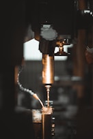 A close-up view of a metalworking machine, focusing on a drill bit actively cutting or drilling into a piece of metal. Sparks and metal shavings are visible, with a flexible coolant hose in the frame. The lighting is dim with warm tones, emphasizing the metallic surfaces.