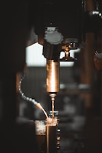 A close-up view of a metalworking machine, focusing on a drill bit actively cutting or drilling into a piece of metal. Sparks and metal shavings are visible, with a flexible coolant hose in the frame. The lighting is dim with warm tones, emphasizing the metallic surfaces.