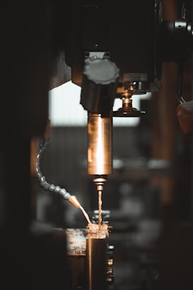A close-up view of a metalworking machine, focusing on a drill bit actively cutting or drilling into a piece of metal. Sparks and metal shavings are visible, with a flexible coolant hose in the frame. The lighting is dim with warm tones, emphasizing the metallic surfaces.