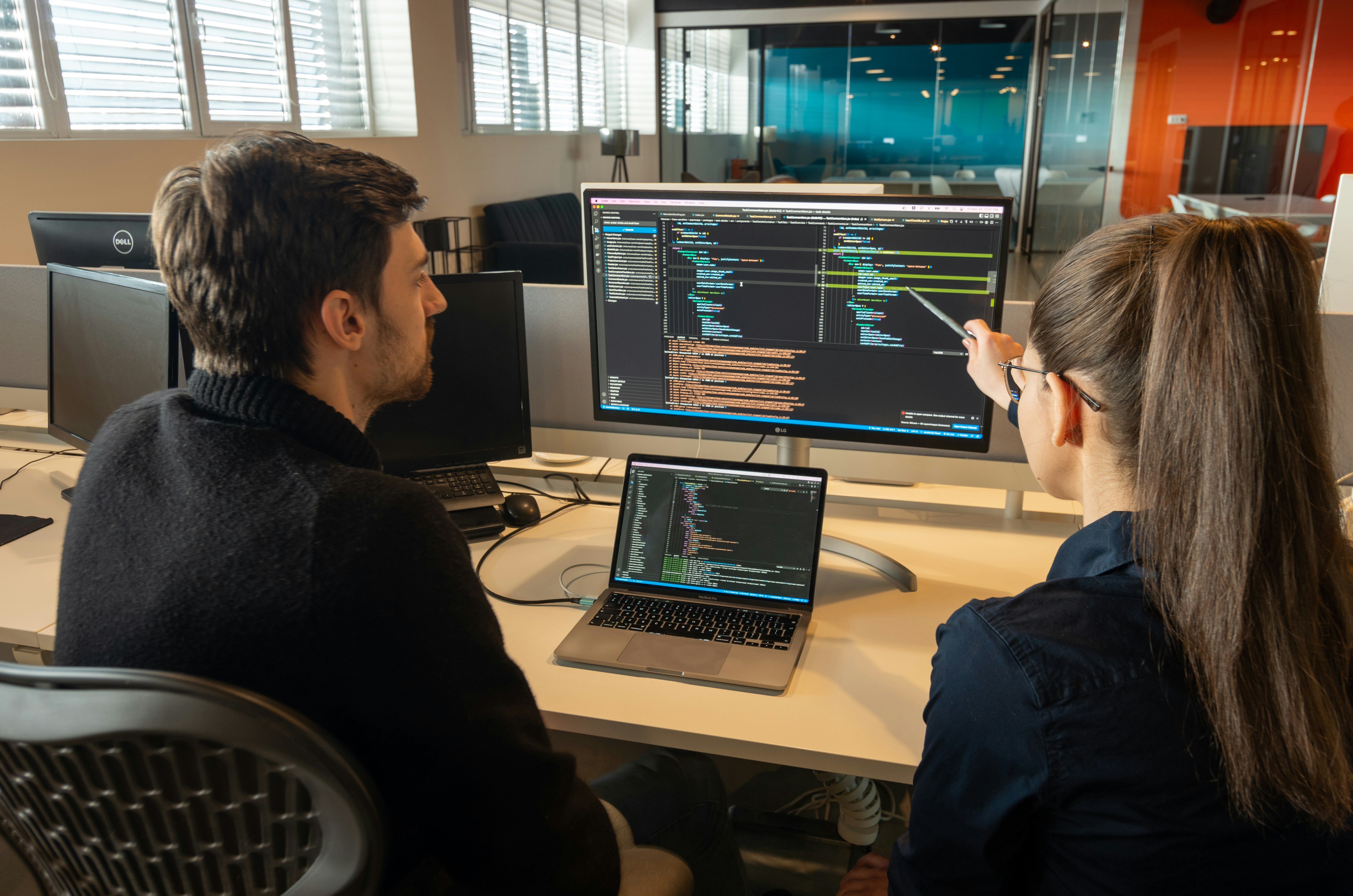 TechVault team members collaborating in modern office space, engaged in animated discussion around a large monitor displaying code and design mockups