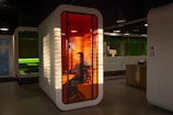 A modern office setting features a small, enclosed booth with a translucent orange interior and a person seated inside using a computer. The booth is illuminated by sunlight filtering through blinds, creating distinct patterns of light and shadow. In the background, another booth with a green interior is visible, and the overall ambiance suggests a futuristic and high-tech environment.