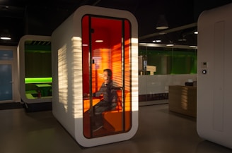 A modern office setting features a small, enclosed booth with a translucent orange interior and a person seated inside using a computer. The booth is illuminated by sunlight filtering through blinds, creating distinct patterns of light and shadow. In the background, another booth with a green interior is visible, and the overall ambiance suggests a futuristic and high-tech environment.