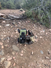 Two remote-controlled toy trucks, one green and one beige, are positioned on a rocky dirt terrain. The trucks appear to be engaged in an off-road activity, with one truck partially climbing over the other. Surrounding the vehicles, the landscape is scattered with stones and dry vegetation, with evergreen trees in the background.