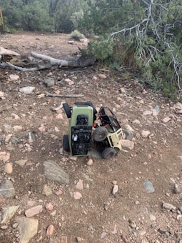 Two remote-controlled toy trucks, one green and one beige, are positioned on a rocky dirt terrain. The trucks appear to be engaged in an off-road activity, with one truck partially climbing over the other. Surrounding the vehicles, the landscape is scattered with stones and dry vegetation, with evergreen trees in the background.