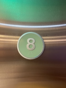 A button labeled with the number 8 is prominently displayed against a metallic background. The button is round with a light green face and a silvery border. Below the number, small raised dots represent Braille symbols.
