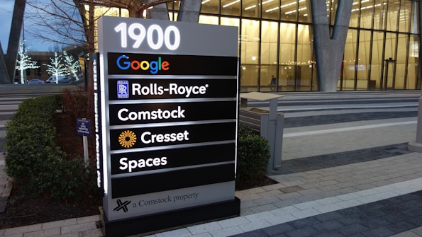 A large directory sign is situated outdoors listing several company names, including Google, Rolls-Royce, Comstock, Cresset, and Spaces, in front of a modern glass building. The ground is paved with tiles, and there are some small shrubs and decorative lights on trees visible in the background.
