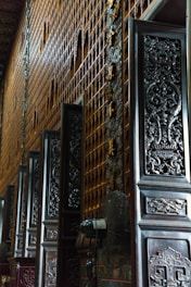 Intricately carved wooden doors line a wall with golden ornamentation in a dimly lit interior space. The details on the doors feature elaborate designs and patterns, contributing to an ornate and historic atmosphere.