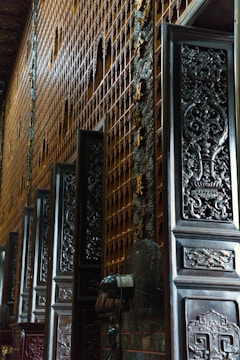 Intricately carved wooden doors line a wall with golden ornamentation in a dimly lit interior space. The details on the doors feature elaborate designs and patterns, contributing to an ornate and historic atmosphere.