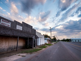 A deserted street with an old building labeled 'Baker's Auction' and several power lines against a backdrop of a cloudy blue sky. The building appears aged and boarded up, suggesting abandonment or disuse. The street is empty, adding to the sense of solitude and stillness.