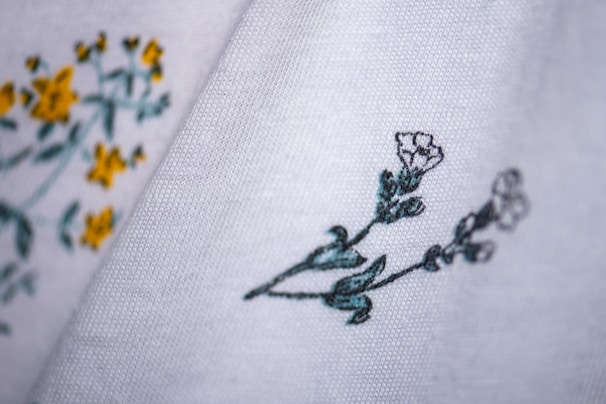 Close-up of fabric with delicate floral embroidery. The fabric is white with embroidered yellow and green flowers on the left side, and green and white flowers on the right side.