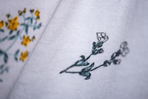 Close-up of fabric with delicate floral embroidery. The fabric is white with embroidered yellow and green flowers on the left side, and green and white flowers on the right side.