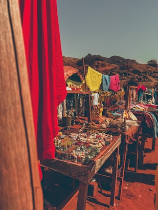 A vibrant outdoor market scene with tables filled with colorful textiles, jewelry, and small glass jars. Brightly colored fabrics are draped on makeshift stands, and the setting is sunlit with a backdrop of arid hills and a clear sky.