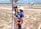 A worker in blue and orange safety gear is climbing an electrical pole using a ladder. They are wearing a white helmet and gloves while holding onto the pole. The background shows a grassy area with a few palm trees, a road, and a distant industrial building under a blue sky with clouds.