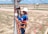 A worker in blue and orange safety gear is climbing an electrical pole using a ladder. They are wearing a white helmet and gloves while holding onto the pole. The background shows a grassy area with a few palm trees, a road, and a distant industrial building under a blue sky with clouds.
