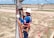 A worker in blue and orange safety gear is climbing an electrical pole using a ladder. They are wearing a white helmet and gloves while holding onto the pole. The background shows a grassy area with a few palm trees, a road, and a distant industrial building under a blue sky with clouds.