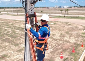 A worker in blue and orange safety gear is climbing an electrical pole using a ladder. They are wearing a white helmet and gloves while holding onto the pole. The background shows a grassy area with a few palm trees, a road, and a distant industrial building under a blue sky with clouds.