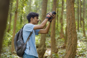 A person with a backpack holds a camera while standing in a forest, surrounded by tall trees and lush greenery. The individual appears focused and intent on capturing a photograph, with dappled sunlight filtering through the foliage.
