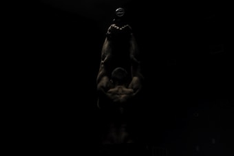 A muscular individual is lifting a dumbbell overhead in a dimly lit environment, highlighting the contours of their muscles. The light source emphasizes the upper body, creating a dramatic shadow effect.