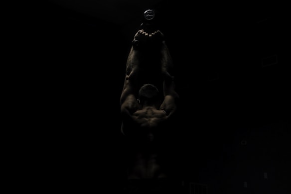 A muscular individual is lifting a dumbbell overhead in a dimly lit environment, highlighting the contours of their muscles. The light source emphasizes the upper body, creating a dramatic shadow effect.