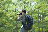 A person holding a camera, poised to take a photograph, is seen against a backdrop of lush green trees. The individual is wearing a casual outfit and a large backpack.