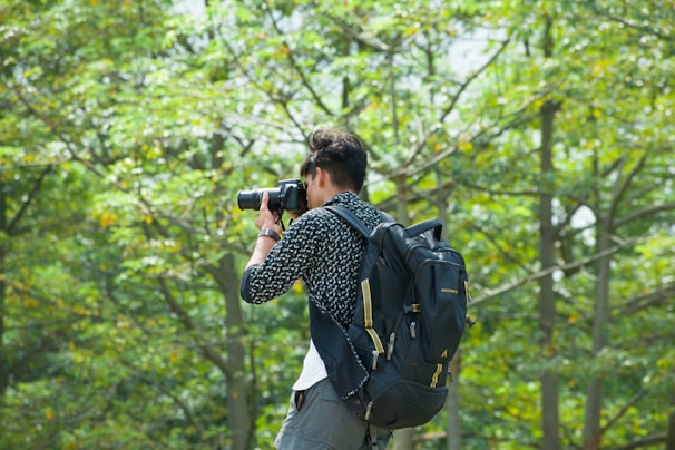 A person holding a camera, poised to take a photograph, is seen against a backdrop of lush green trees. The individual is wearing a casual outfit and a large backpack.