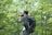 A person holding a camera, poised to take a photograph, is seen against a backdrop of lush green trees. The individual is wearing a casual outfit and a large backpack.