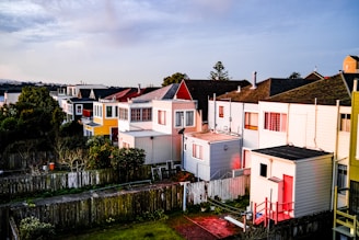 A row of colorful, two-story houses with varying shades of red, yellow, and white are lined up along a narrow street. The houses display a mix of modern and traditional styles, with well-maintained exteriors. A fence runs parallel to the houses, bordering a grassy area. Trees and shrubs partially obscure the houses, and a water body is visible in the background.