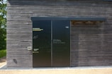 A dark wooden wall with a large black sliding door is seen. The wall is composed of horizontal wooden planks with visible screws. The door has a matte finish with various inscriptions and messages in chalk or paint. To the left of the door, some text is printed in three languages, calling for opinions. Bright sunlight casts shadows on the surface, and there is greenery peeking from the side.