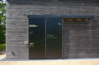 A dark wooden wall with a large black sliding door is seen. The wall is composed of horizontal wooden planks with visible screws. The door has a matte finish with various inscriptions and messages in chalk or paint. To the left of the door, some text is printed in three languages, calling for opinions. Bright sunlight casts shadows on the surface, and there is greenery peeking from the side.
