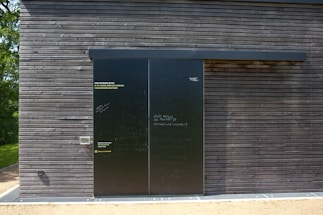 A dark wooden wall with a large black sliding door is seen. The wall is composed of horizontal wooden planks with visible screws. The door has a matte finish with various inscriptions and messages in chalk or paint. To the left of the door, some text is printed in three languages, calling for opinions. Bright sunlight casts shadows on the surface, and there is greenery peeking from the side.