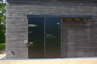 A dark wooden wall with a large black sliding door is seen. The wall is composed of horizontal wooden planks with visible screws. The door has a matte finish with various inscriptions and messages in chalk or paint. To the left of the door, some text is printed in three languages, calling for opinions. Bright sunlight casts shadows on the surface, and there is greenery peeking from the side.