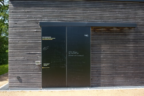 A dark wooden wall with a large black sliding door is seen. The wall is composed of horizontal wooden planks with visible screws. The door has a matte finish with various inscriptions and messages in chalk or paint. To the left of the door, some text is printed in three languages, calling for opinions. Bright sunlight casts shadows on the surface, and there is greenery peeking from the side.