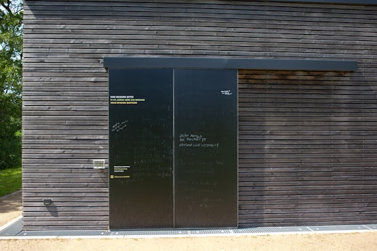 A dark wooden wall with a large black sliding door is seen. The wall is composed of horizontal wooden planks with visible screws. The door has a matte finish with various inscriptions and messages in chalk or paint. To the left of the door, some text is printed in three languages, calling for opinions. Bright sunlight casts shadows on the surface, and there is greenery peeking from the side.