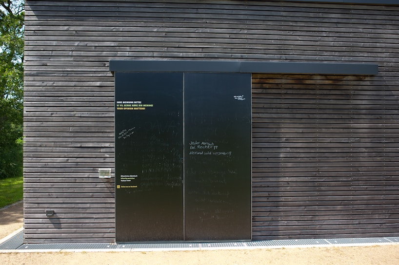 A dark wooden wall with a large black sliding door is seen. The wall is composed of horizontal wooden planks with visible screws. The door has a matte finish with various inscriptions and messages in chalk or paint. To the left of the door, some text is printed in three languages, calling for opinions. Bright sunlight casts shadows on the surface, and there is greenery peeking from the side.