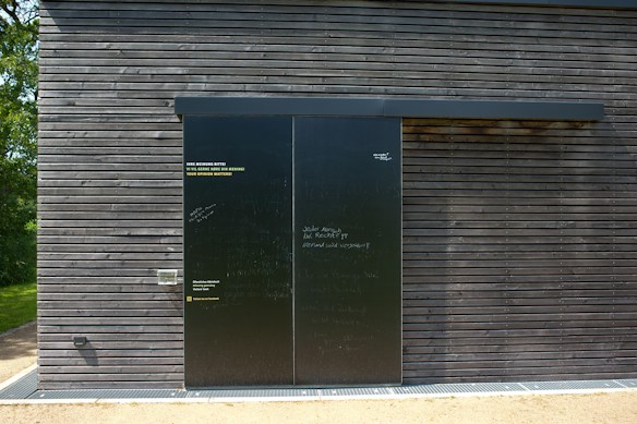 A dark wooden wall with a large black sliding door is seen. The wall is composed of horizontal wooden planks with visible screws. The door has a matte finish with various inscriptions and messages in chalk or paint. To the left of the door, some text is printed in three languages, calling for opinions. Bright sunlight casts shadows on the surface, and there is greenery peeking from the side.