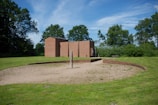 A modern brick building is set amidst a lush green landscape. In front of the building, a circular sandy area is bordered by a low brick wall. There is a solitary vertical wooden post placed centrally within the sandy circle. Trees and bushes are scattered around the area, providing a backdrop of greenery under a clear blue sky.