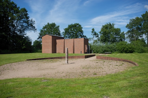 A modern brick building is set amidst a lush green landscape. In front of the building, a circular sandy area is bordered by a low brick wall. There is a solitary vertical wooden post placed centrally within the sandy circle. Trees and bushes are scattered around the area, providing a backdrop of greenery under a clear blue sky.